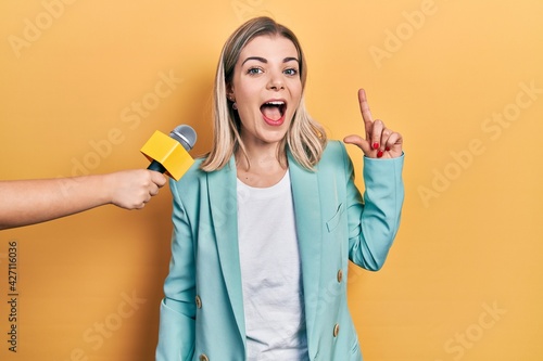 Beautiful caucasian woman being interviewed by reporter holding microphone pointing finger up with successful idea. exited and happy. number one.