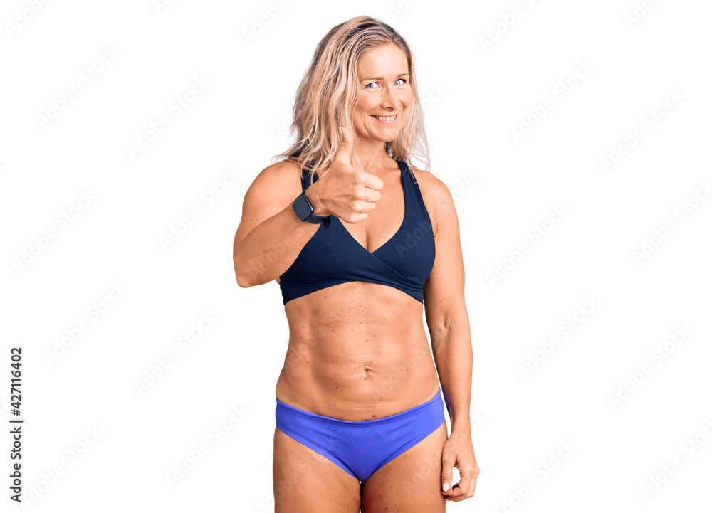 Middle age fit blonde woman wearing bikini doing happy thumbs up gesture with hand. approving expression looking at the camera showing success. Middle age fit blonde woman wearing bikini doing happy thumbs up gesture with hand. approving expression looking at the camera showing success.