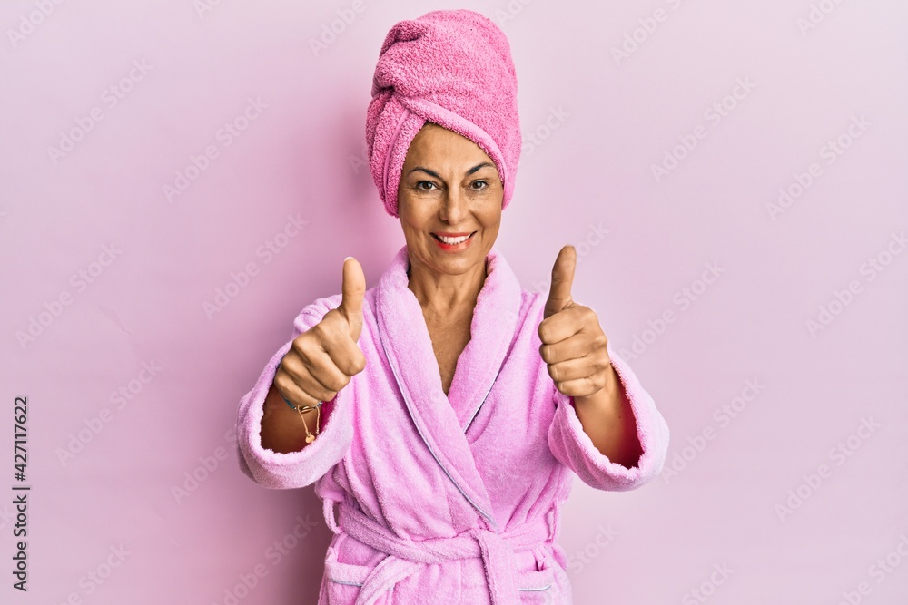Middle age hispanic woman wearing shower towel cap and bathrobe approving doing positive gesture with hand, thumbs up smiling and happy for success. winner gesture. Middle age hispanic woman wearing shower towel cap and bathrobe approving doing positive gesture with hand, thumbs up smiling and happy for success. winner gesture.