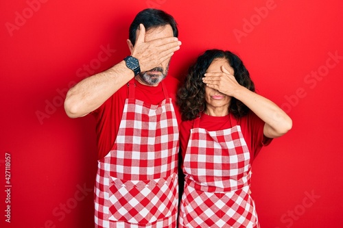 Middle age couple of hispanic woman and man wearing cook apron covering eyes with hand, looking serious and sad. sightless, hiding and rejection concept