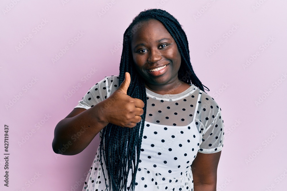 Young black woman with braids wearing casual clothes doing happy thumbs up gesture with hand. approving expression looking at the camera showing success. Young black woman with braids wearing casual clothes doing happy thumbs up gesture with hand. approving expression looking at the camera showing success.