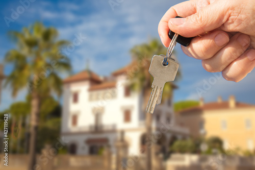 Hand with a key is on the background of a luxury house with palm trees. Selective focus on the key. The concept of selling expensive housing.