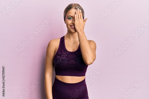 Beautiful blonde woman wearing sportswear over pink background covering one eye with hand  confident smile on face and surprise emotion.