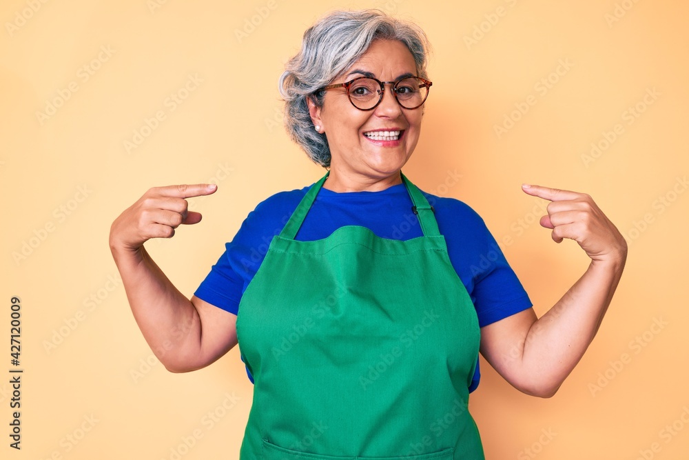Senior hispanic woman wearing apron and glasses looking confident with smile on face, pointing oneself with fingers proud and happy.