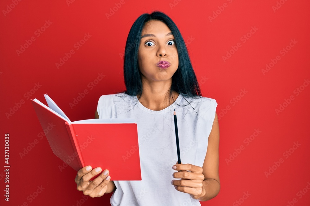 Young african american woman holding book and pencil puffing cheeks with funny face. mouth inflated with air, catching air. Young african american woman holding book and pencil puffing cheeks with funny face. mouth inflated with air, catching air.