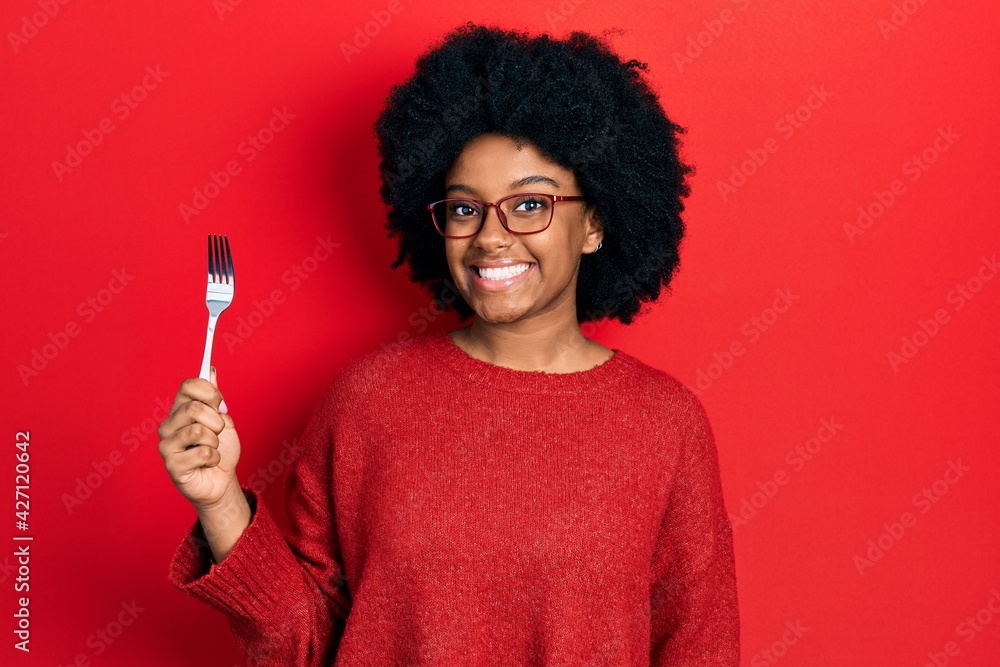 Young african american woman holding one silver fork looking positive and happy standing and smiling with a confident smile showing teeth Young african american woman holding one silver fork looking positive and happy standing and smiling with a confident smile showing teeth