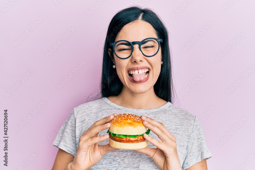 Beautiful young woman eating tasty hamburger sticking tongue out happy with funny expression. Beautiful young woman eating tasty hamburger sticking tongue out happy with funny expression.