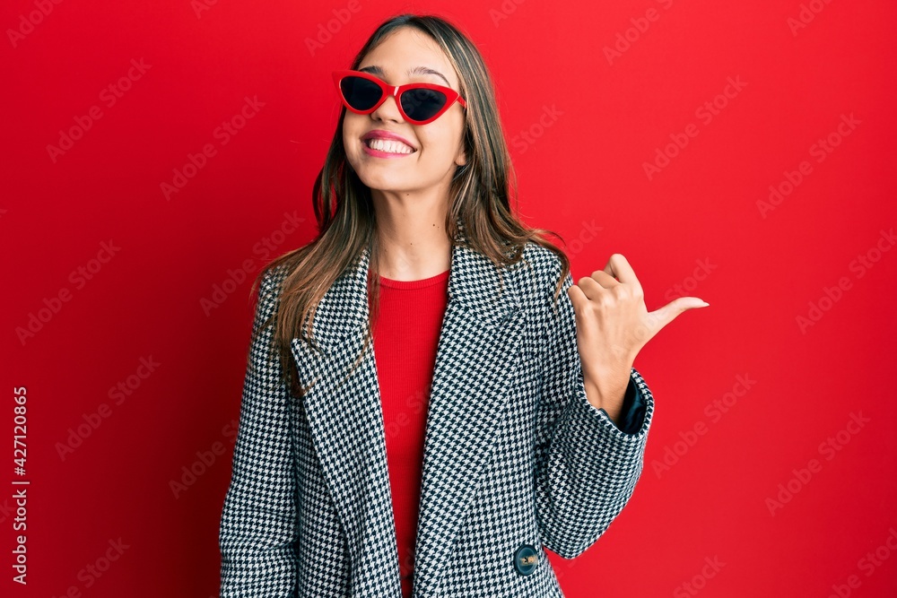 Young brunette woman wearing fashion and modern look smiling with happy face looking and pointing to the side with thumb up. Young brunette woman wearing fashion and modern look smiling with happy face looking and pointing to the side with thumb up.