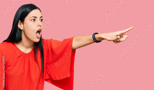 Beautiful young woman wearing casual clothes pointing with finger surprised ahead, open mouth amazed expression, something on the front © Krakenimages.com