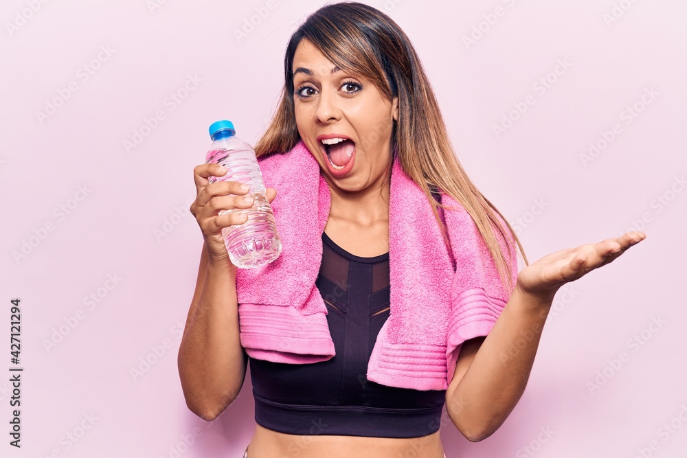 Young beautiful woman wearing sportswear and towel holding bottle of water celebrating achievement with happy smile and winner expression with raised hand Young beautiful woman wearing sportswear and towel holding bottle of water celebrating achievement with happy smile and winner expression with raised hand