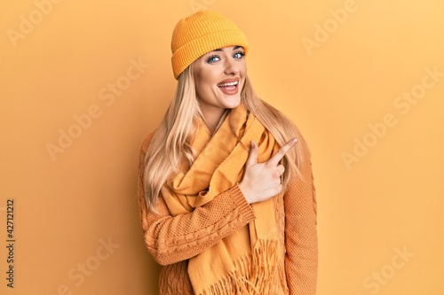 Young caucasian woman wearing wool winter sweater and cap cheerful with a smile of face pointing with hand and finger up to the side with happy and natural expression on face © Krakenimages.com
