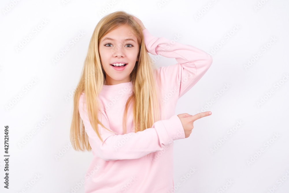 beautiful caucasian little girl wearing pink hoodie over white background feeling positive has amazed expression, indicates something. One hand on head and pointing with other hand. beautiful caucasian little girl wearing pink hoodie over white background feeling positive has amazed expression, indicates something. One hand on head and pointing with other hand.