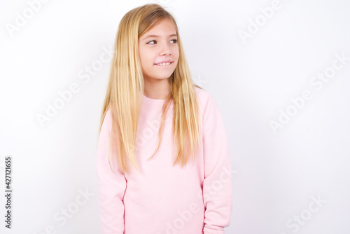 beautiful caucasian little girl wearing pink hoodie over white background with thoughtful expression, looks away keeps hands down bitting his lip thinks about something pleasant. © Jihan