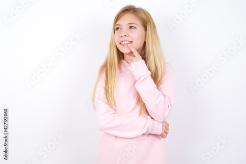 beautiful caucasian little girl wearing pink hoodie over white background with thoughtful expression  looks to the camera  keeps hand near face  bitting a finger thinks about something pleasant.