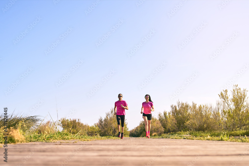 Active couple running during a warm summer day. Wearing pink shirts and short pants. Both are wearing sunglasses. Running towards the camera. Low angle shot. Active couple running during a warm summer day. Wearing pink shirts and short pants. Both are wearing sunglasses. Running towards the camera. Low angle shot.