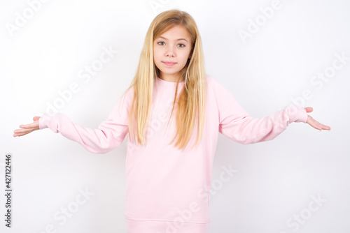 Puzzled and clueless beautiful caucasian little girl wearing pink hoodie over white background with arms out, shrugging shoulders, saying: who cares, so what, I don't know. Negative human emotions. © Jihan
