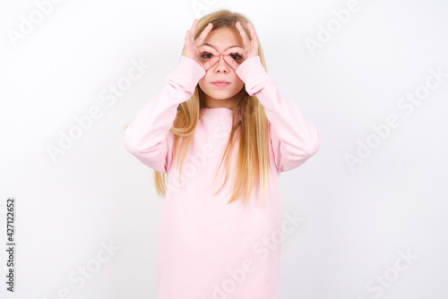Playful excited beautiful caucasian little girl wearing pink hoodie over white background showing Ok sign with both hands on eyes, pretending to wear spectacles.