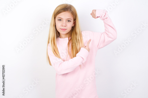 Smiling beautiful caucasian little girl wearing pink hoodie over white background raises hand to show muscles, feels confident in victory, strong and independent.