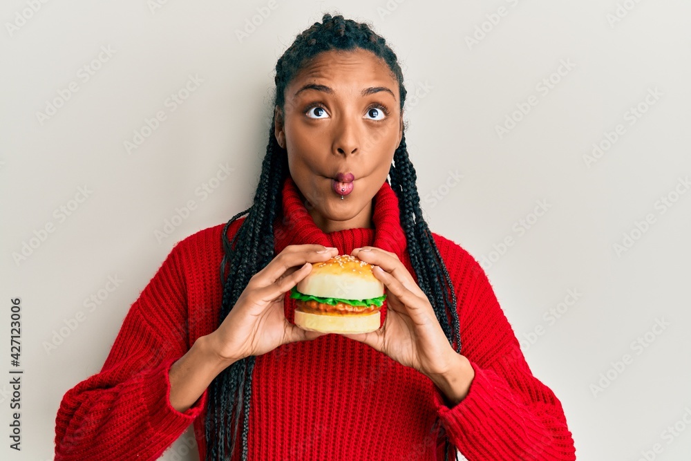 African american woman with braids eating hamburger making fish face with  mouth and squinting eyes, crazy and comical. Stock Photo