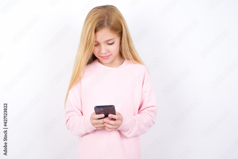 Smiling beautiful caucasian little girl wearing pink hoodie over white back using cell phone, messaging, being happy to text with friends, looking at smartphone. Modern technologies and communication. Smiling beautiful caucasian little girl wearing pink hoodie over white back using cell phone, messaging, being happy to text with friends, looking at smartphone. Modern technologies and communication.