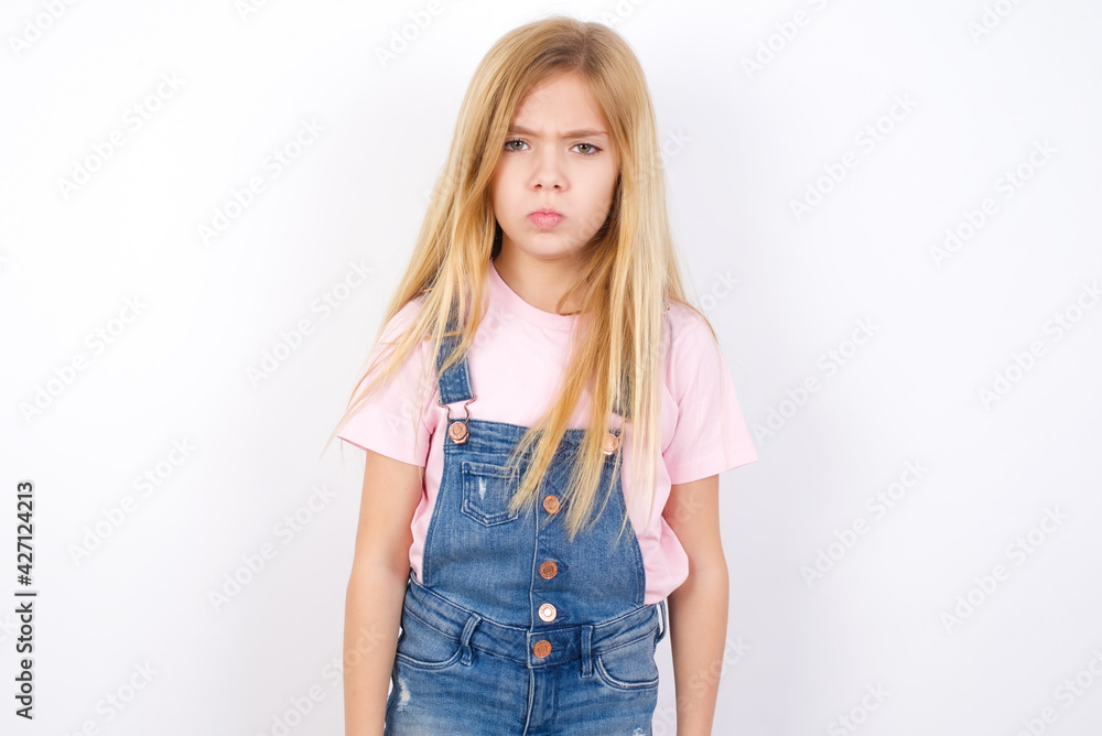 Obraz premium Offended dissatisfied beautiful caucasian little girl wearing jeans overall over white background with moody displeased expression at camera being disappointed by something