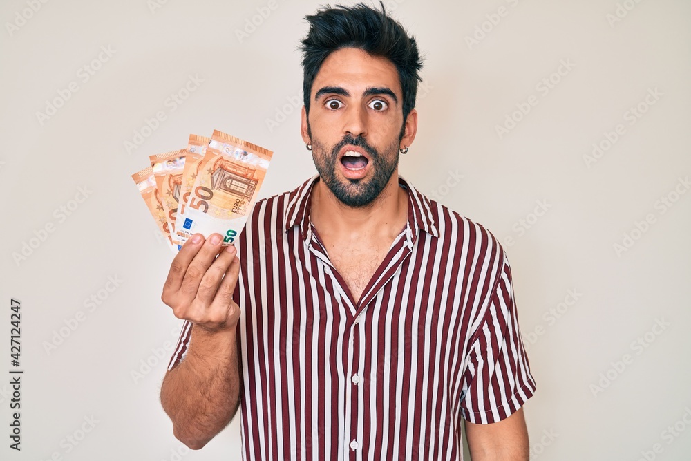 Handsome hispanic man with beard holding euro banknotes scared and amazed with open mouth for surprise, disbelief face Handsome hispanic man with beard holding euro banknotes scared and amazed with open mouth for surprise, disbelief face