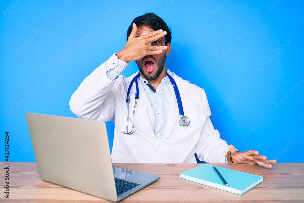 Handsome hispanic man wearing doctor uniform working at the clinic peeking in shock covering face and eyes with hand, looking through fingers afraid Handsome hispanic man wearing doctor uniform working at the clinic peeking in shock covering face and eyes with hand, looking through fingers afraid