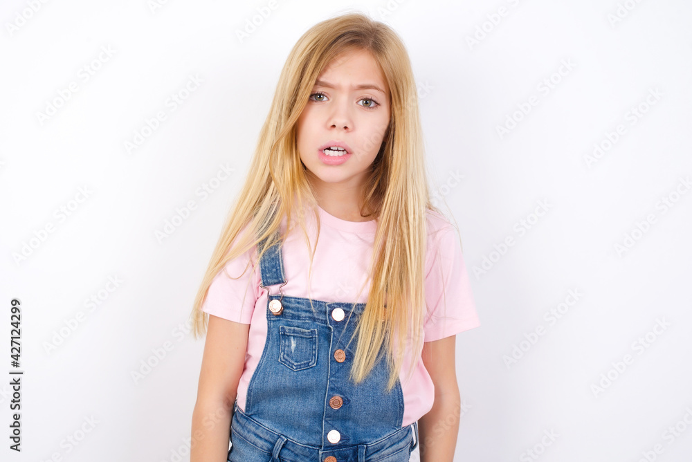 Obraz premium Portrait of dissatisfied beautiful caucasian little girl wearing jeans overall over white wall smirks face, purses lips and looks with annoyance at camera, discontent hearing something unpleasant