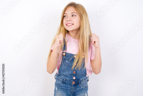 beautiful caucasian little girl wearing jeans overall over white background clenches fists and awaits for something nice happened looks away bites lips and waits announcement of results