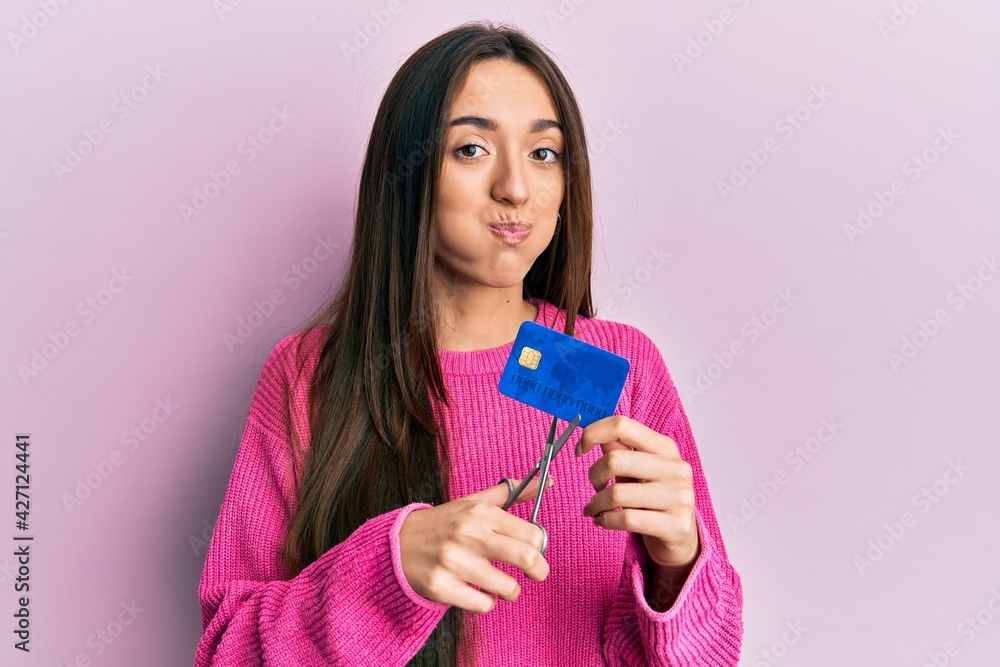 Young hispanic girl cutting credit card using scissors puffing cheeks with funny face. mouth inflated with air, catching air. Young hispanic girl cutting credit card using scissors puffing cheeks with funny face. mouth inflated with air, catching air.