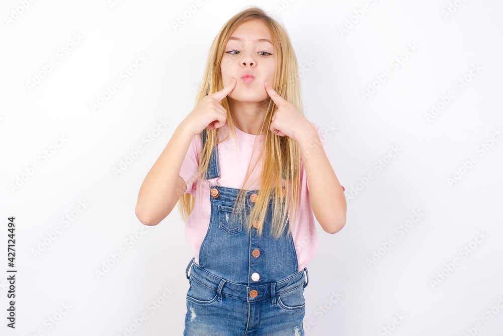 beautiful caucasian little girl wearing jeans overall over white background crosses eyes and makes fish lips funny grimace beautiful caucasian little girl wearing jeans overall over white background crosses eyes and makes fish lips funny grimace