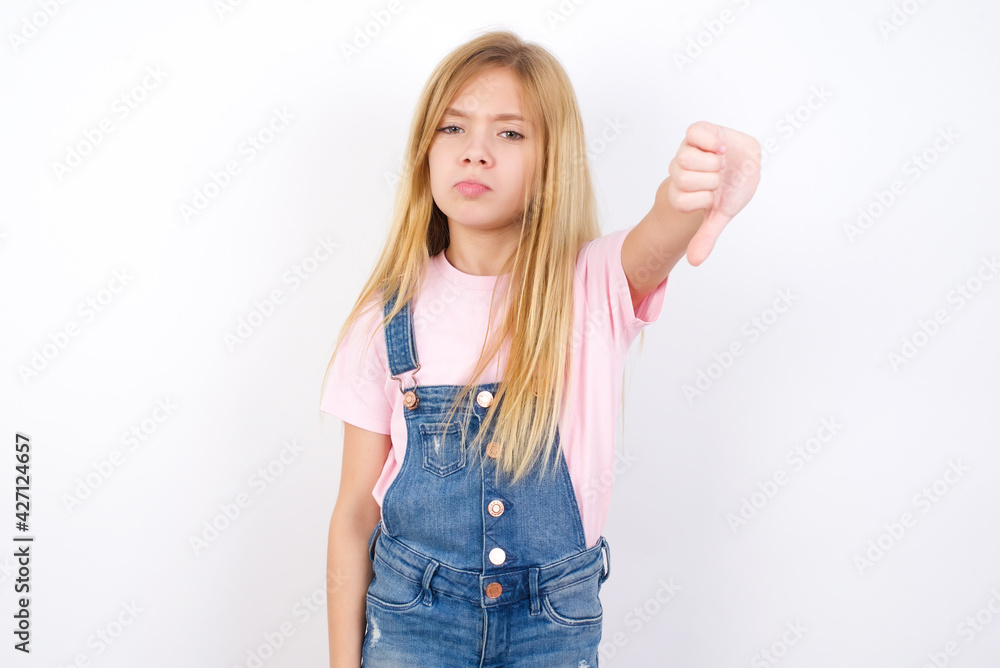beautiful caucasian little girl wearing jeans overall over white background looking unhappy and angry showing rejection and negative with thumbs down gesture. Bad expression. beautiful caucasian little girl wearing jeans overall over white background looking unhappy and angry showing rejection and negative with thumbs down gesture. Bad expression.