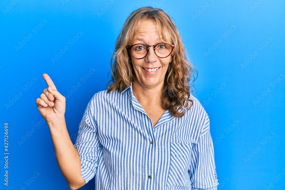 Middle age blonde woman wearing casual clothes and glasses with a big smile on face, pointing with hand finger to the side looking at the camera. Middle age blonde woman wearing casual clothes and glasses with a big smile on face, pointing with hand finger to the side looking at the camera.