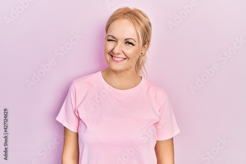 Young blonde woman wearing casual pink t shirt winking looking at the camera with sexy expression, cheerful and happy face.