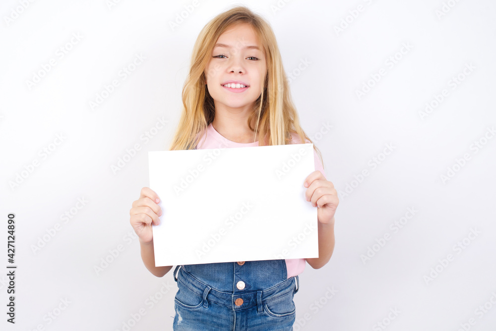Happy beautiful caucasian little girl wearing jeans overall over white background Holding Empty Paper Board Advertising Offer Text Standing. Autumn Advertisement Banner. Happy beautiful caucasian little girl wearing jeans overall over white background Holding Empty Paper Board Advertising Offer Text Standing. Autumn Advertisement Banner.