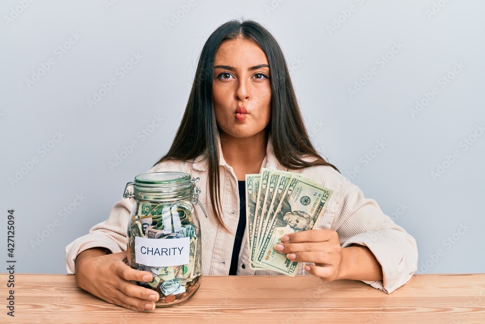 Beautiful hispanic woman holding charity jar with dollars making fish face with mouth and squinting eyes, crazy and comical. Beautiful hispanic woman holding charity jar with dollars making fish face with mouth and squinting eyes, crazy and comical.