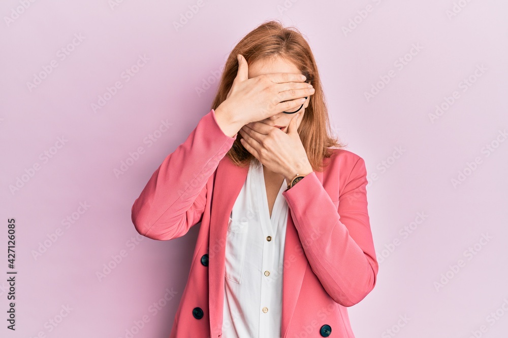 Young caucasian woman wearing business style and glasses covering eyes and mouth with hands, surprised and shocked. hiding emotion Young caucasian woman wearing business style and glasses covering eyes and mouth with hands, surprised and shocked. hiding emotion