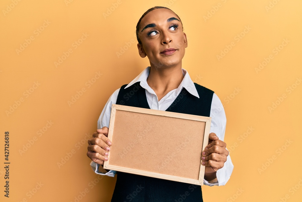 Hispanic man wearing make up and long hair holding empty corkboard smiling looking to the side and staring away thinking. Hispanic man wearing make up and long hair holding empty corkboard smiling looking to the side and staring away thinking.