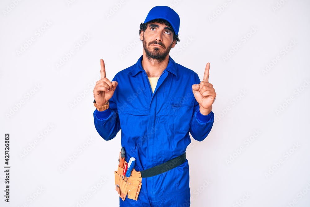 Handsome young man with curly hair and bear weaing handyman uniform pointing up looking sad and upset, indicating direction with fingers, unhappy and depressed.
