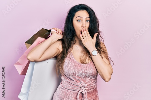 Young hispanic woman holding shopping bags covering mouth with hand, shocked and afraid for mistake. surprised expression