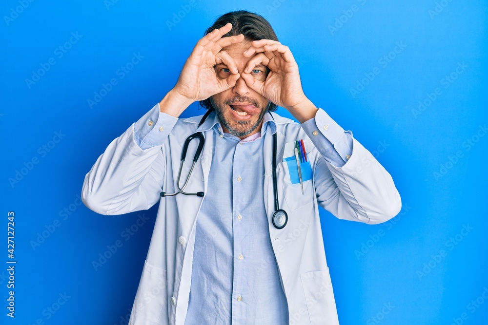 Middle age handsome man wearing doctor uniform and stethoscope doing ok gesture like binoculars sticking tongue out, eyes looking through fingers. crazy expression.