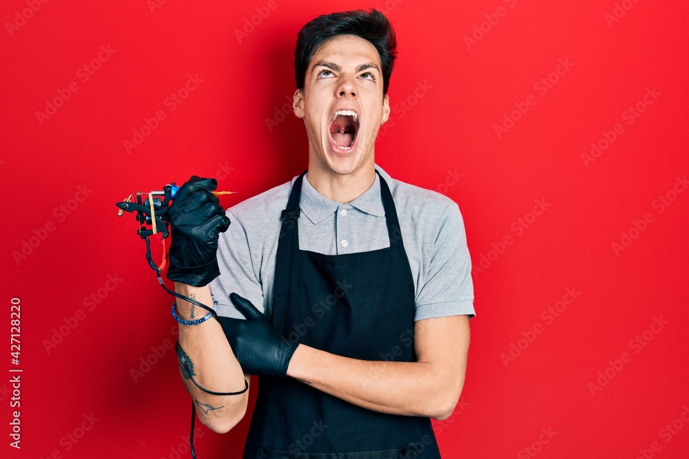 Young hispanic man tattoo artist wearing professional uniform and gloves holding tattooer machine annoyed and frustrated shouting with anger, yelling crazy with anger and hand raised Young hispanic man tattoo artist wearing professional uniform and gloves holding tattooer machine annoyed and frustrated shouting with anger, yelling crazy with anger and hand raised