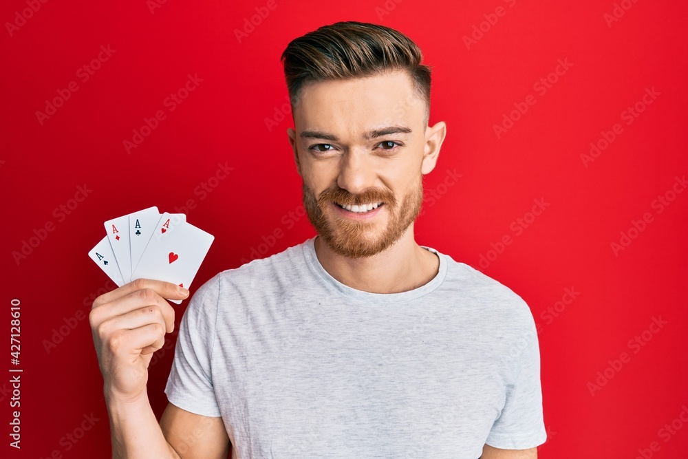 Young redhead man holding ace poker cards looking positive and happy standing and smiling with a confident smile showing teeth Young redhead man holding ace poker cards looking positive and happy standing and smiling with a confident smile showing teeth