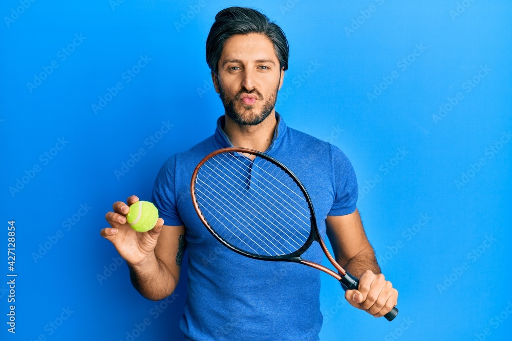 Young hispanic man playing tennis holding racket and ball looking at the camera blowing a kiss being lovely and sexy. love expression.