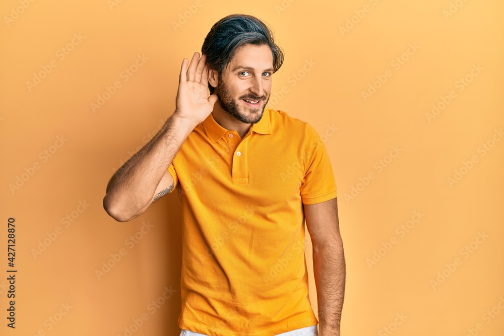 Young hispanic man wearing casual yellow t shirt smiling with hand over ear listening an hearing to rumor or gossip. deafness concept. Young hispanic man wearing casual yellow t shirt smiling with hand over ear listening an hearing to rumor or gossip. deafness concept.