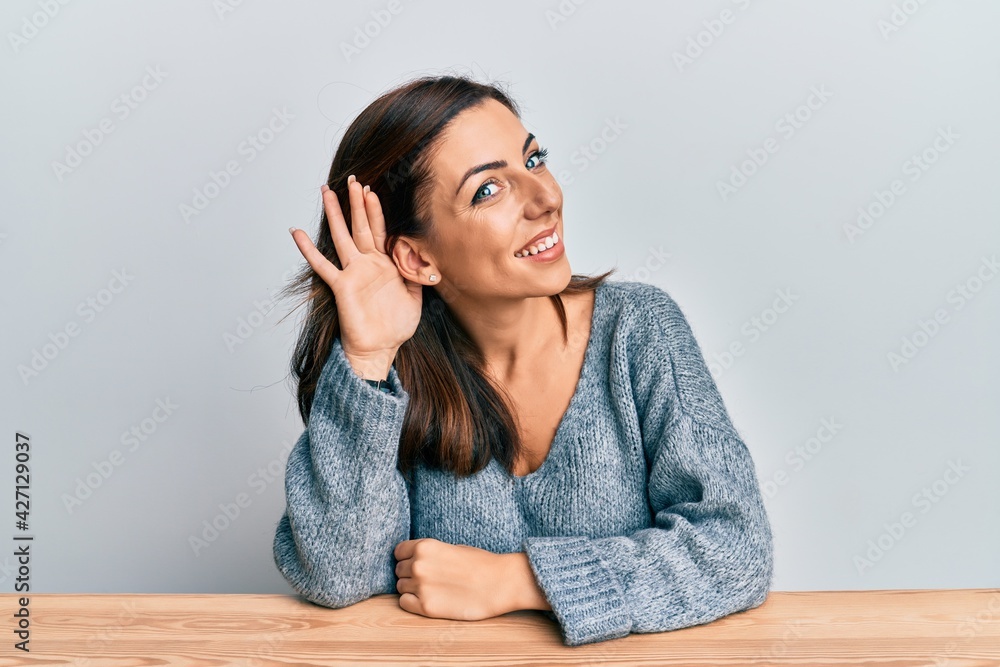 Young brunette woman wearing casual sweater sitting on the table smiling with hand over ear listening and hearing to rumor or gossip. deafness concept. Young brunette woman wearing casual sweater sitting on the table smiling with hand over ear listening and hearing to rumor or gossip. deafness concept.