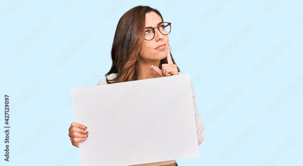 Young brunette woman holding blank empty banner serious face thinking about question with hand on chin, thoughtful about confusing idea Young brunette woman holding blank empty banner serious face thinking about question with hand on chin, thoughtful about confusing idea