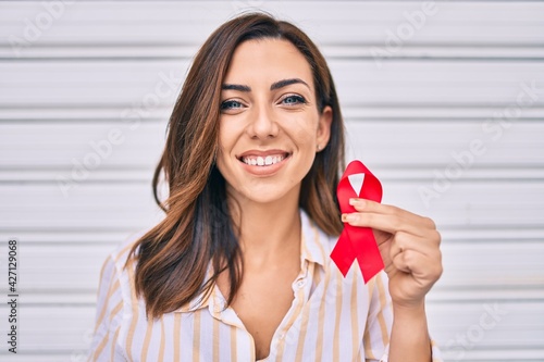 Фотография Young hispanic woman smiling happy holding red hiv ribbon standing at the city