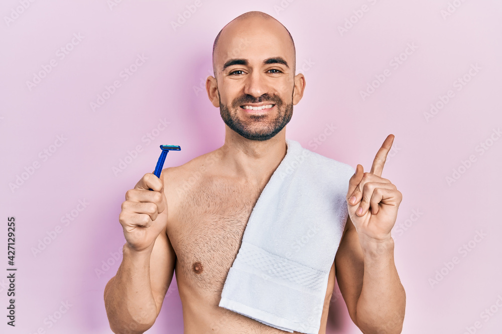 Young bald man shirtless holding razor smiling happy pointing with hand and finger to the side Young bald man shirtless holding razor smiling happy pointing with hand and finger to the side