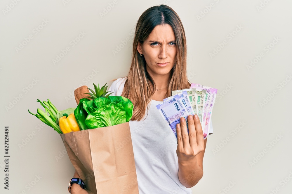 Brunette young woman holding groceries and indian rupee banknotes skeptic and nervous, frowning upset because of problem. negative person. Brunette young woman holding groceries and indian rupee banknotes skeptic and nervous, frowning upset because of problem. negative person.